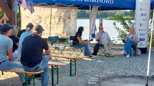 Zdjęcie przedstawiające siedzących na krzesłach uczestników debaty w ramach wydarzenia Tour de Konstytucja Romana Sobotkę, pracownika UODO, Monikę Maćkowiak, sędzię Sądu Rejonowego w Bydgoszczy, i moderatorkę debaty. Nad nimi rozłożony duży parasol z napisem Gmina Koronowo. Na pierwszym planie, po lewej stronie, na długich ławach siedzi publiczność.